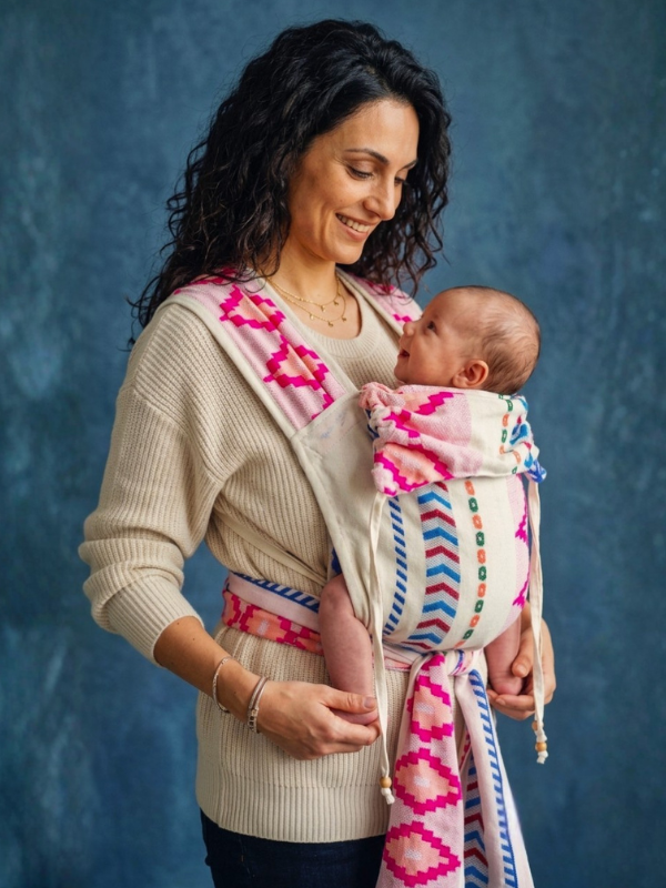 A woman wearing a cream ribbed knit baby carrier wrap with bright pink, blue, and orange geometric patterns, holding a baby against a textured blue studio backdrop.