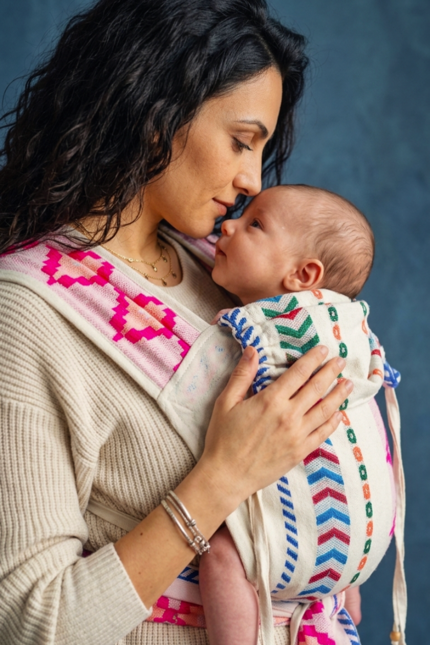 A woman wearing a cream knit sweater and silver bracelets holds a baby in a woven wrap carrier. The carrier has a white base with colorful geometric patterns in pink, blue, green, and orange.