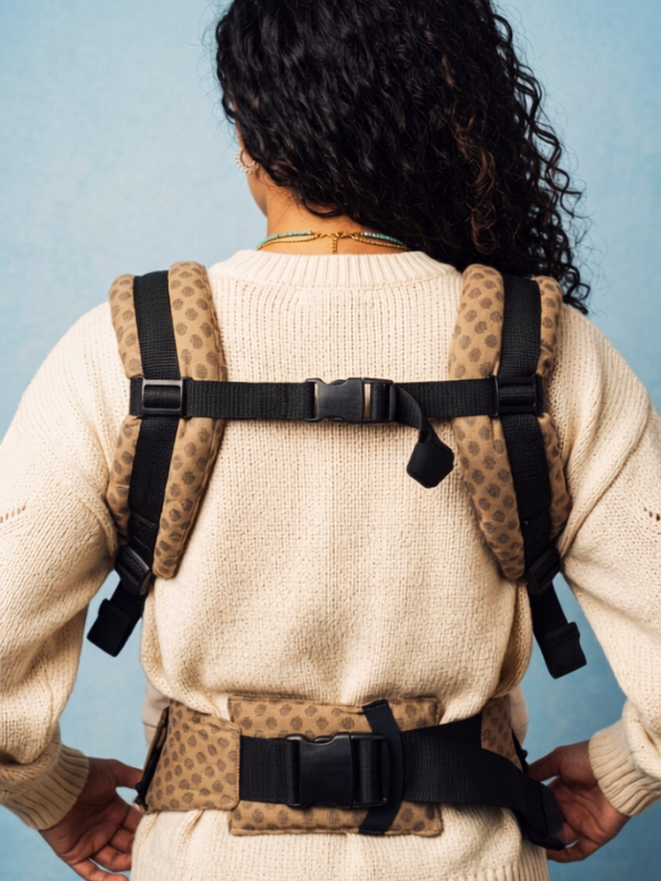 A person viewed from behind wearing a beige knit sweater and a tan-and-black baby support harness with adjustable straps and padded shoulder sections, against a light blue background.
